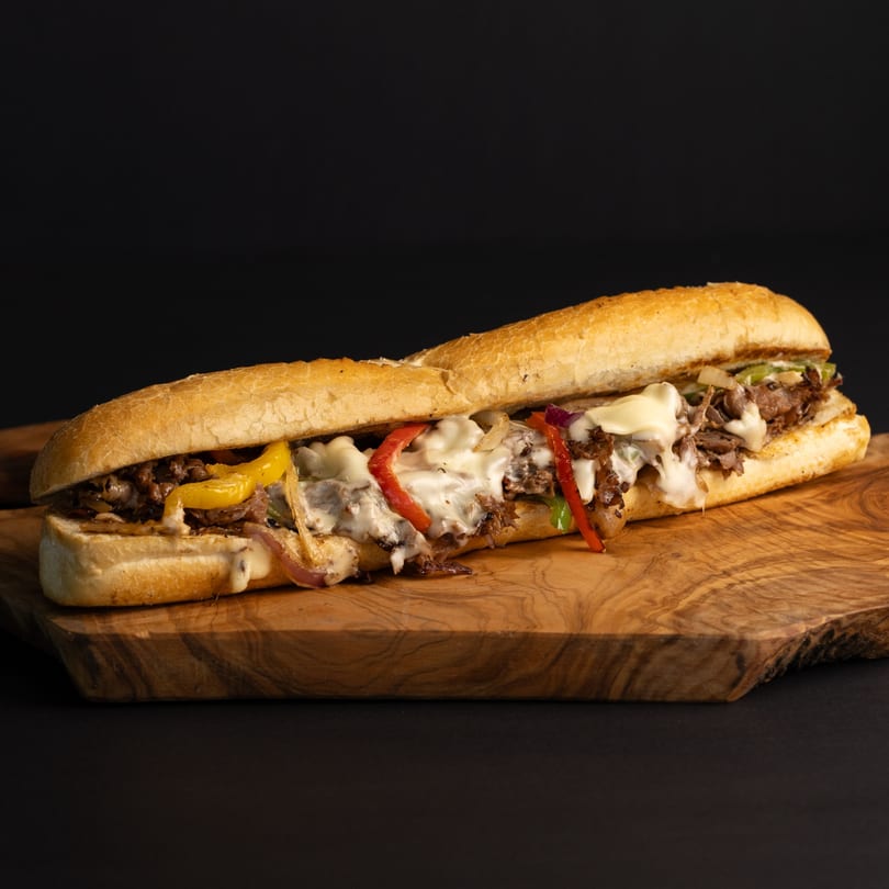 A Dogs & Deli cheesesteak sandwich with melted cheese, steak, and colorful peppers on a wooden board.