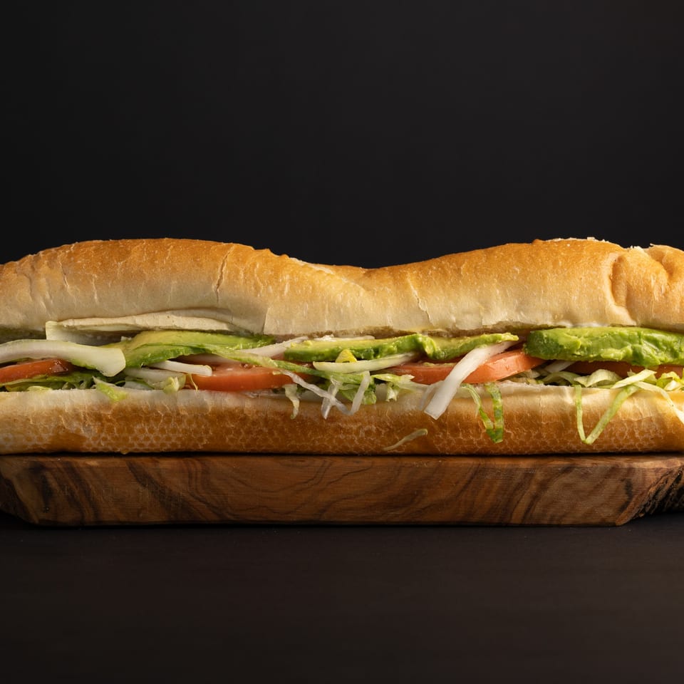 A long deli sandwich with avocado, tomato, lettuce, and onions on a wooden board.