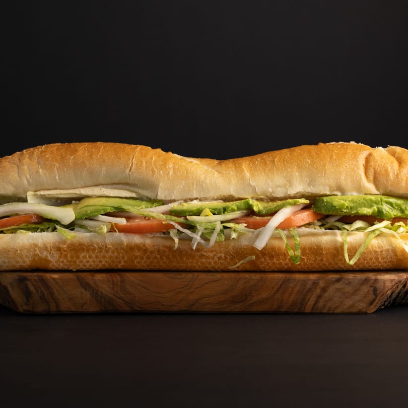 A long deli sandwich with avocado, tomato, lettuce, and onions on a wooden board.