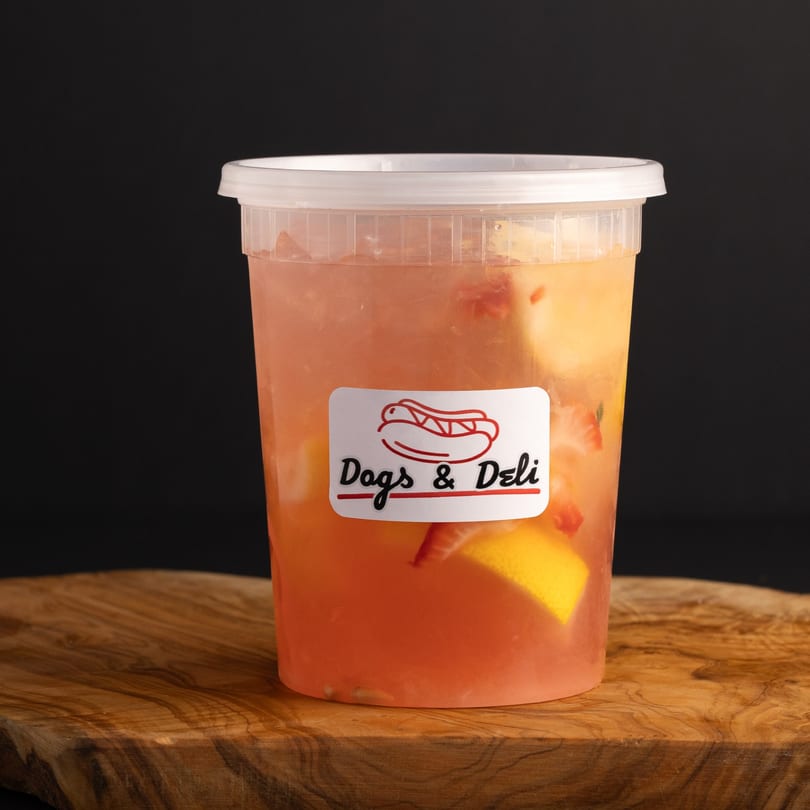 Dogs & Deli branded plastic cup filled with pink lemonade, lemon slices, and strawberries on a wooden board.