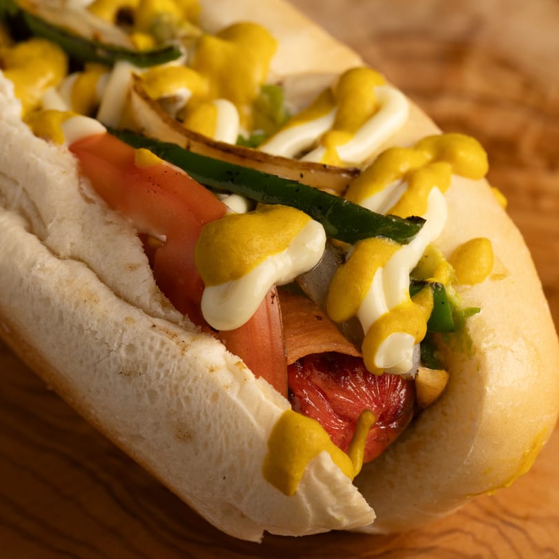 Close-up of a Dogs & Deli hot dog with mustard, mayonnaise, onions, and green peppers.