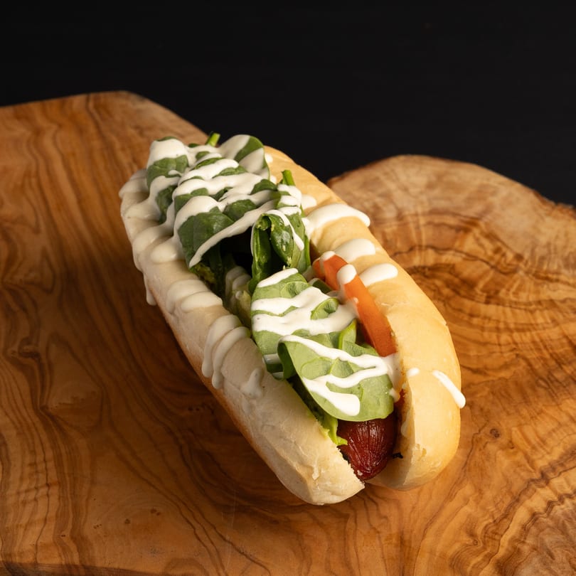 Dogs & Deli hot dog with spinach, carrot, and white sauce on a wooden board.