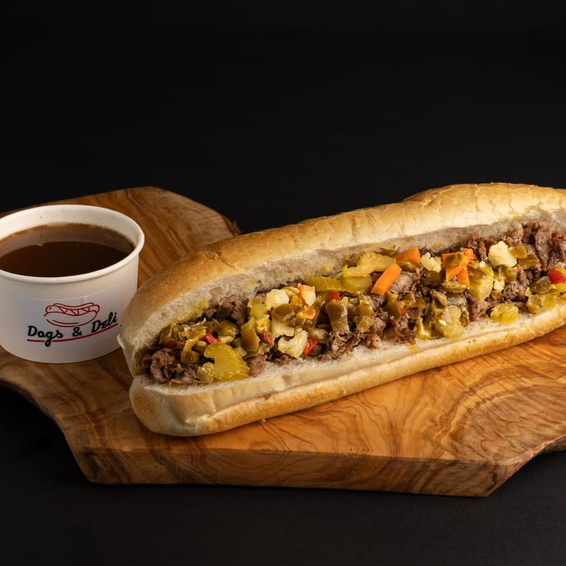 Dogs & Deli's Italian beef sandwich with giardiniera and a side of au jus on a wooden board.