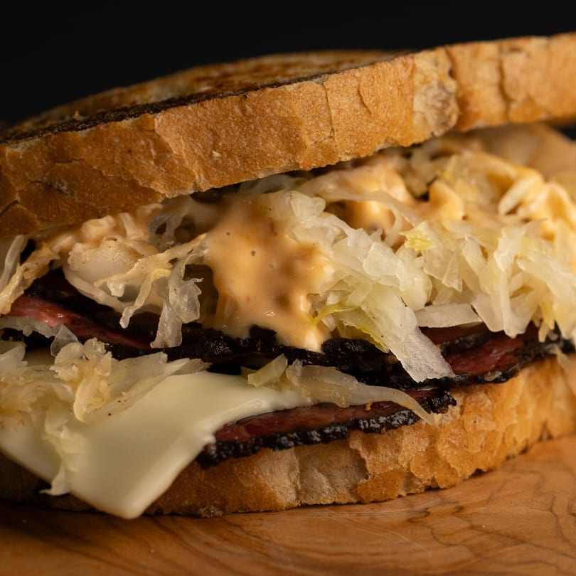 Close-up of a Dogs & Deli Reuben sandwich with pastrami, Swiss cheese, sauerkraut, and Russian dressing.